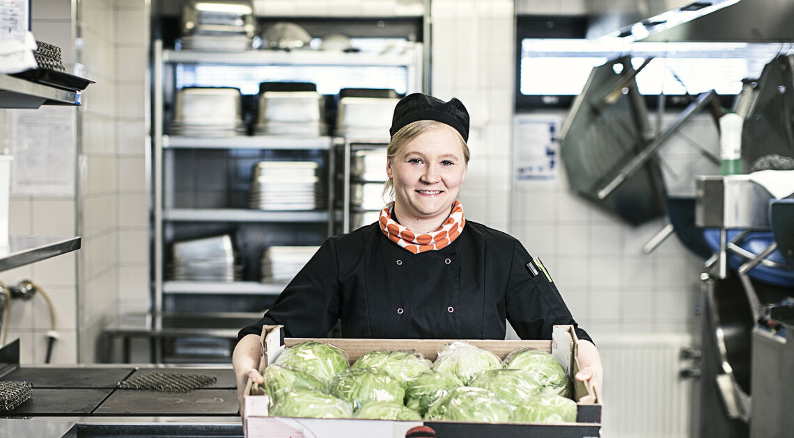 Ruokapalveluvastaava Viivi Tammisto hymyilee työpisteessä keititössä, kädessään hän pitää laatikkoa joka on täynnä tuorettaa salaattia.